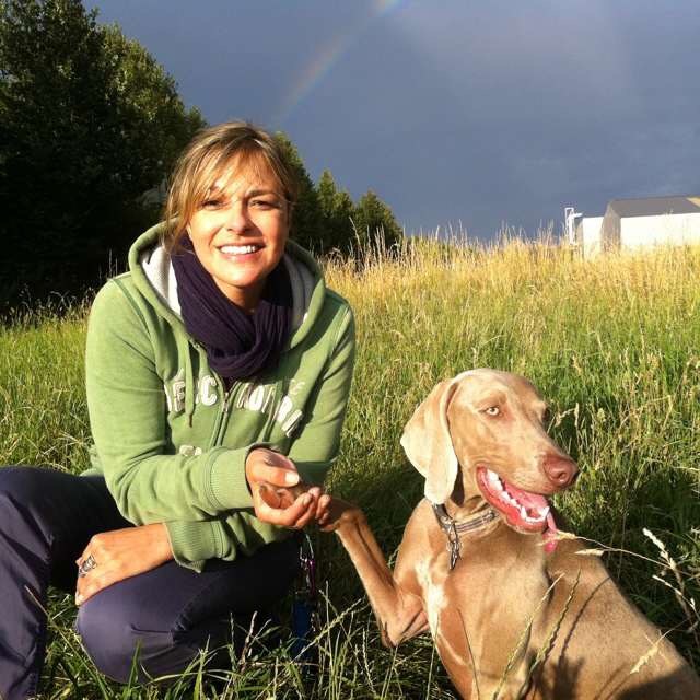 Alice Häuser sitzt mit Maya auf einer Wiese, im Hintergrund ist ein Regenbogen zu sehen.